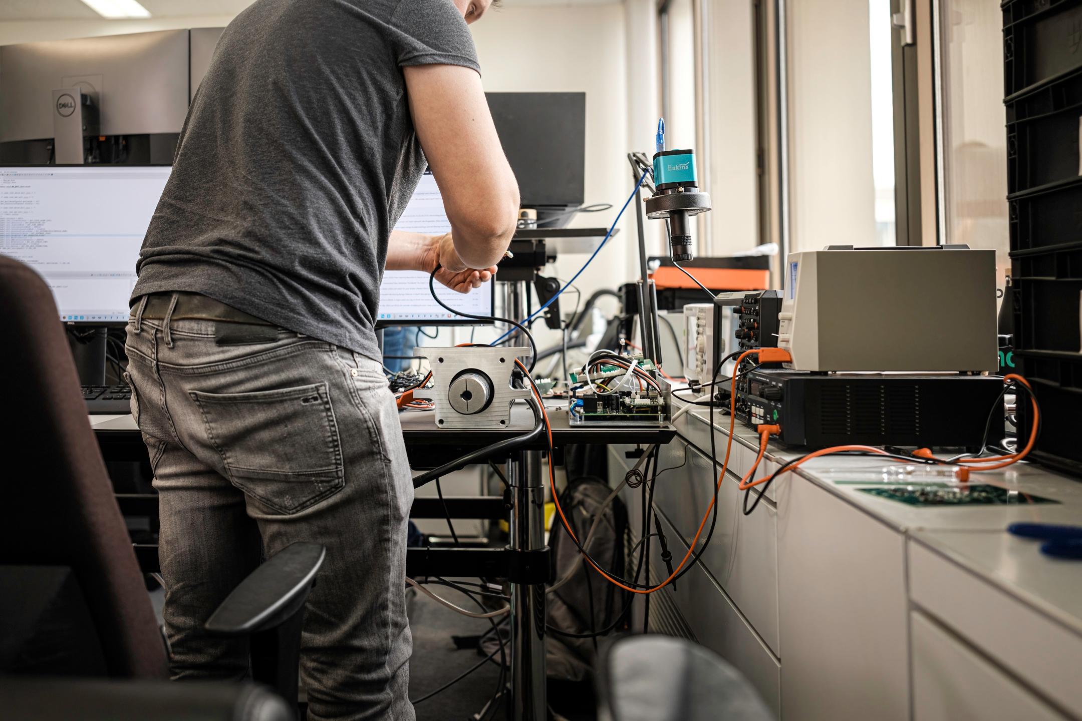 Ein Entwickler am Messplatz von Akrodyn: ein Motor in einer Aluminiumaufnahme, daneben eine offene Leiterplatte, ein Oszilloskop und ein Labornetzteil; auf dem Bildschirm im Hintergrund Quellcode.